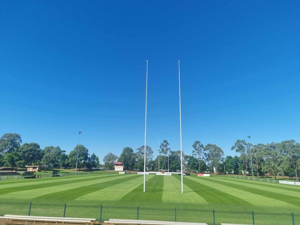 A rugby pitch with green grass, trees in the background and blue skies