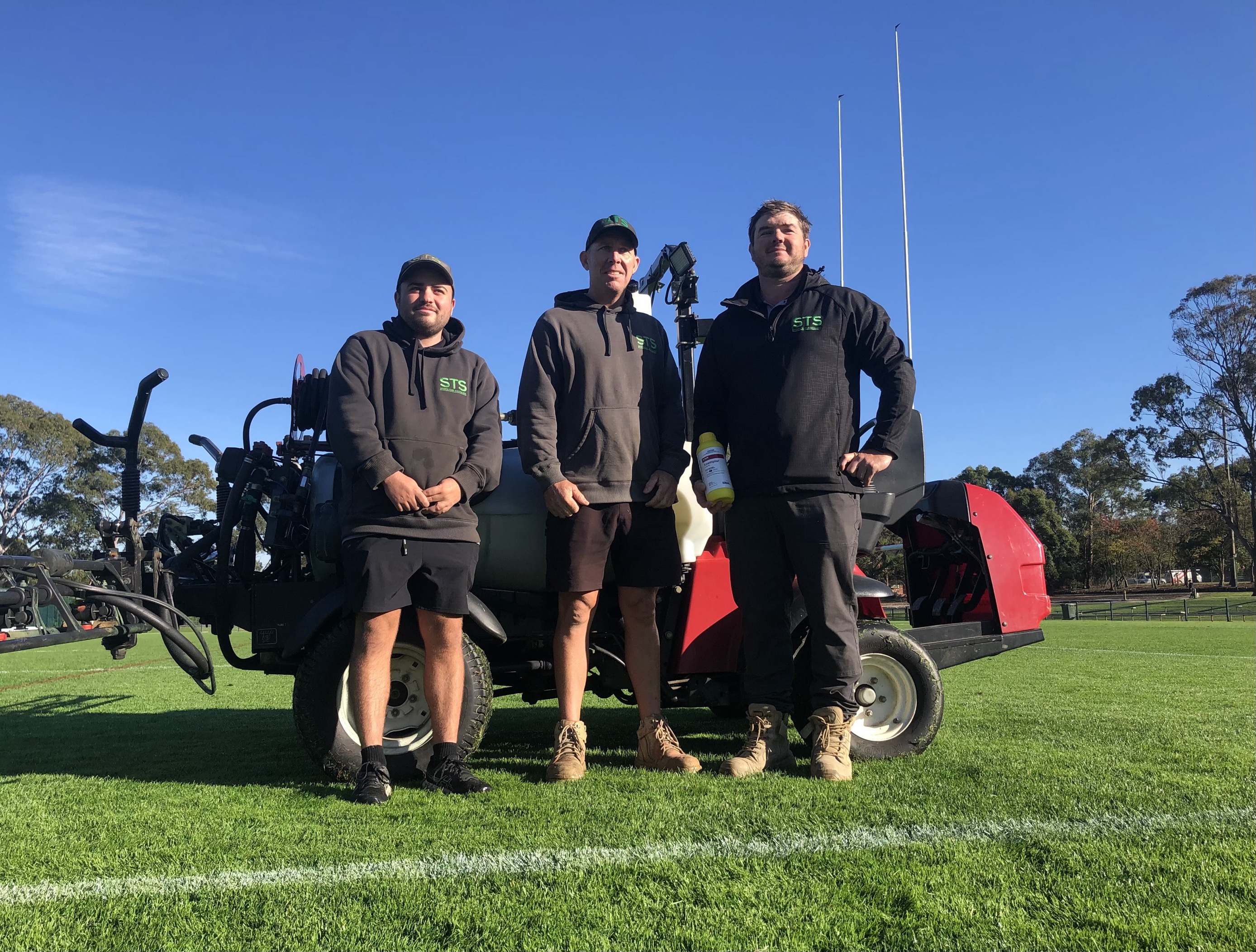 three men stand in front of a machine on a rugby pitch